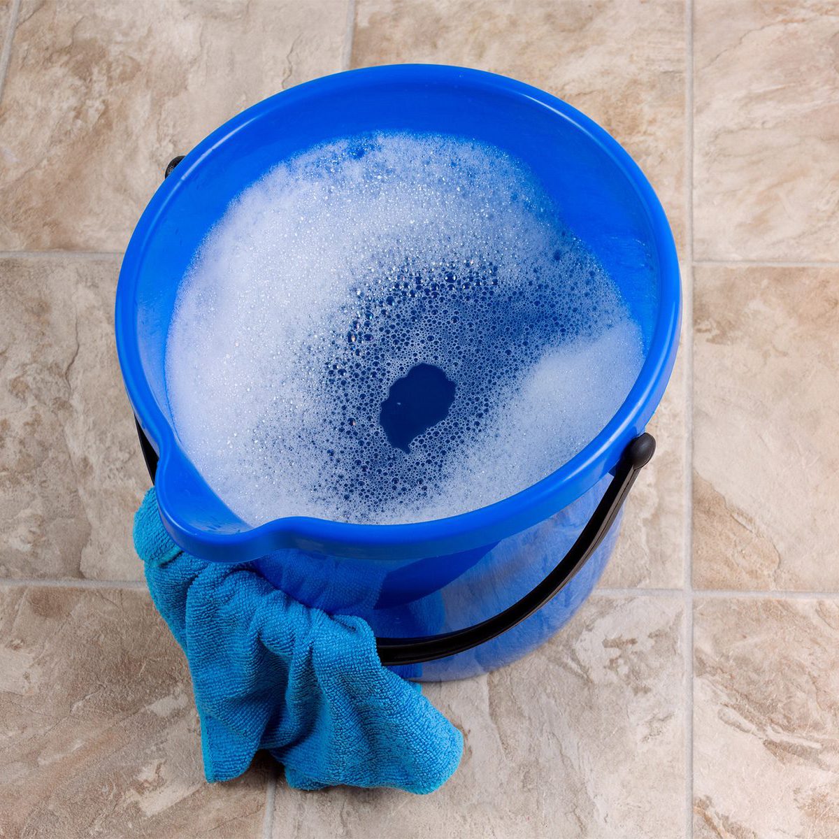 A blue plastic bucket filled with soapy water sits on a tiled floor. A blue cleaning cloth is draped over the edge of the bucket. The surrounding floor tiles have a beige and cream pattern.